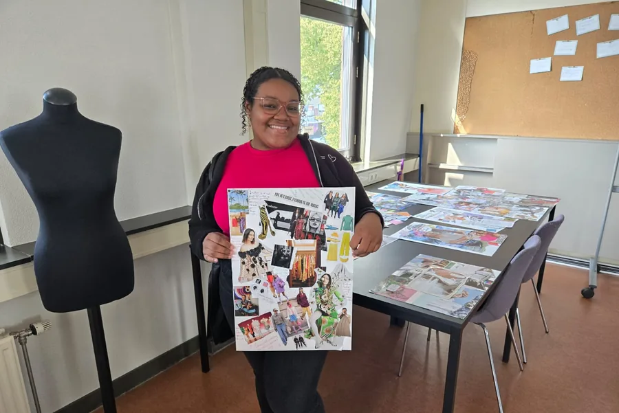 In een naaiatelier houdt iemand een moodboard omhoog met uitgeknipte modebeelden erop geplakt; ernaast staat een paspop en achterin ligt een tafel vol andere bladen.