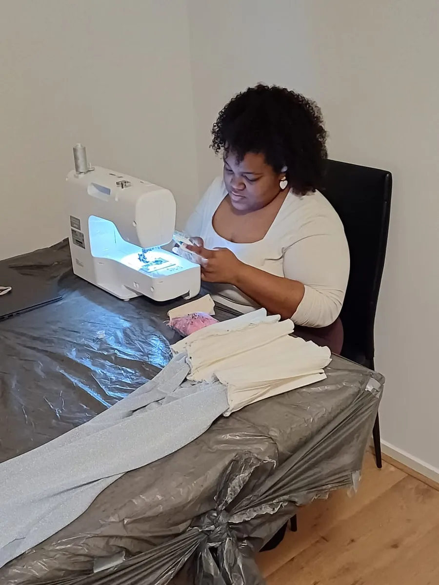 Iemand werkt aan een naaimachine aan een afgedekte tafel, met een stapel geknipte stofdelen binnen handbereik.