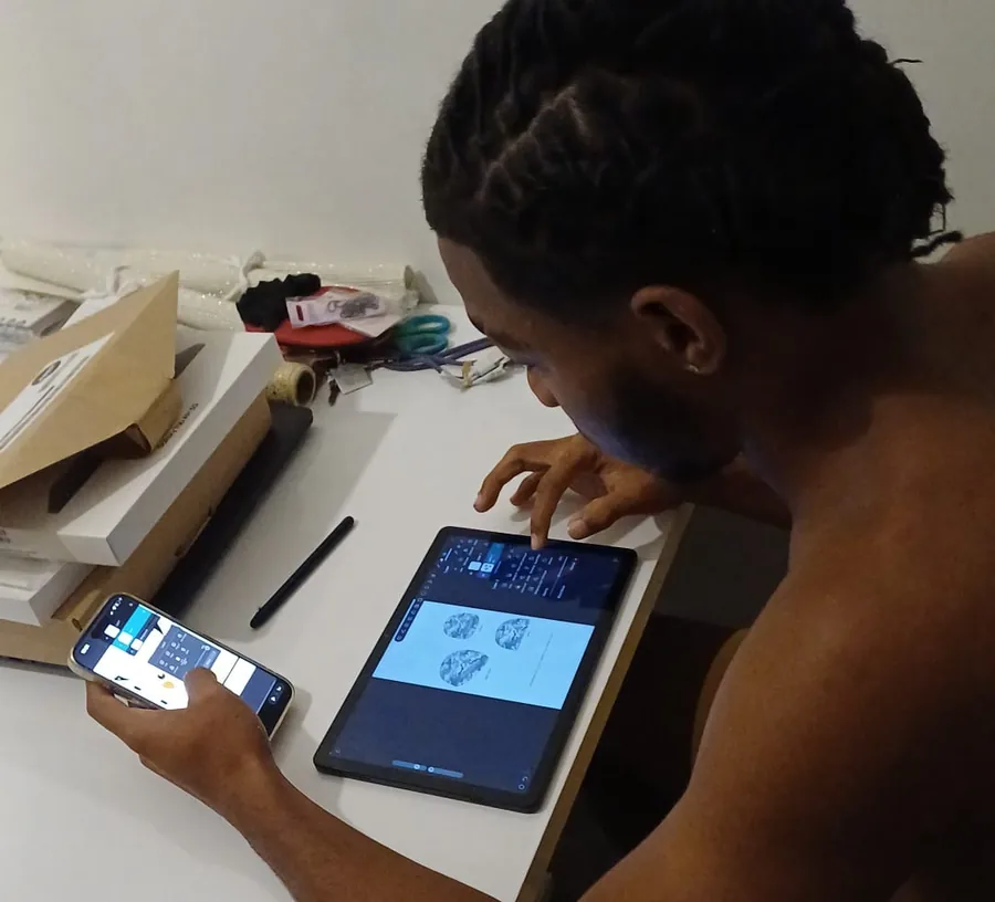 Iemand buigt zich over een werktafel en tekent met een vinger een ontwerp op een tablet; in de andere hand een telefoon met hetzelfde programma, en naast de tablet liggen een stylus, een schaar en garen.