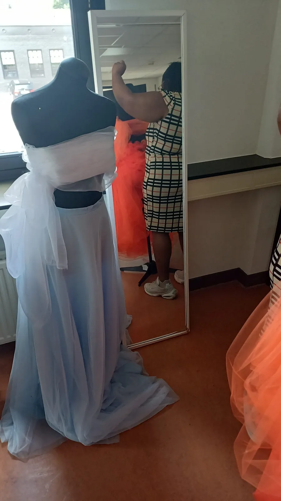 Een lichte japon van tule op een paspop voor het raam, met de oranje rok en een staande spiegel erachter.